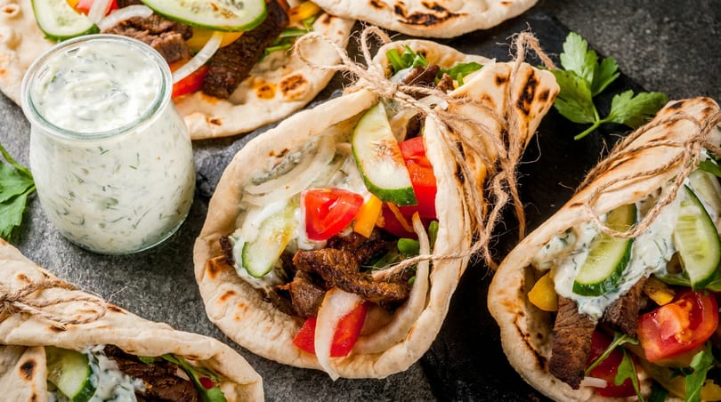 Multiple pita wraps with grilled meat, fresh vegetables, and creamy tzatziki sauce, plus a jar of dip.