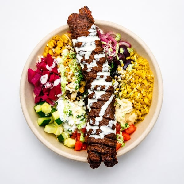 Colorful kebab bowl with meat, white sauce, corn, pickled vegetables, and fresh cucumbers.