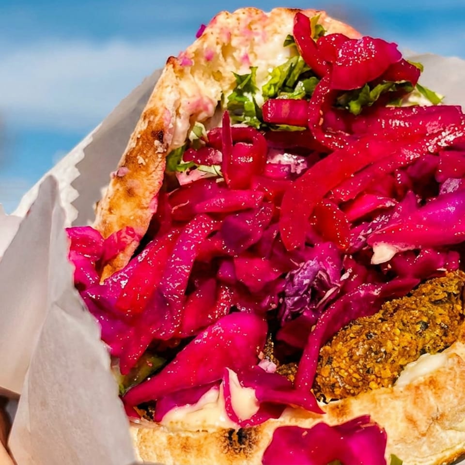 Hand holding a vibrant falafel pita sandwich with bright pink pickled cabbage and sauce outdoors.
