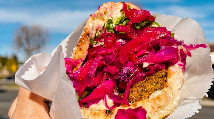 Hand holding a vibrant falafel pita sandwich with bright pink pickled cabbage and sauce outdoors.