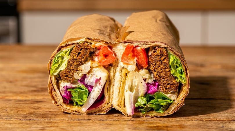 Falafel wrap cut in half, displaying falafel, lettuce, tomatoes, and sauce on a rustic wooden table.