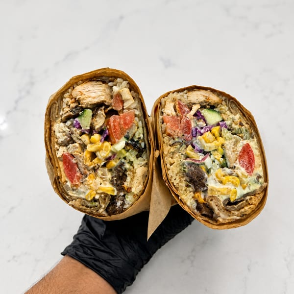 Gloved hand holding a large burrito cut in half, revealing colorful chicken, rice, and vegetable fillings.