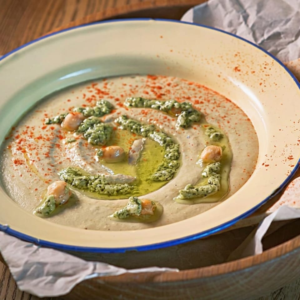 Creamy hummus topped with olive oil, green sauce, chickpeas, paprika, and pita bread.