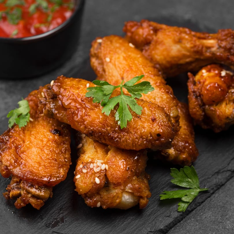 Crispy glazed chicken wings with sesame seeds and parsley, served with a side of red dipping sauce.