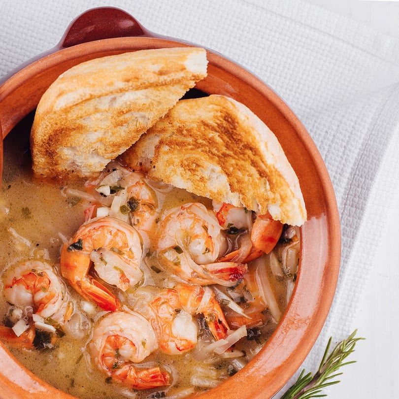 Shrimp scampi in a rustic bowl with toasted bread and rosemary on a white tablecloth.