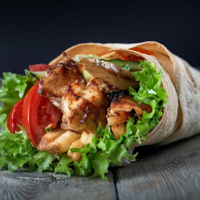 Grilled chicken shawarma wrap with lettuce, tomatoes, cucumbers, and fries on a dark wooden surface.