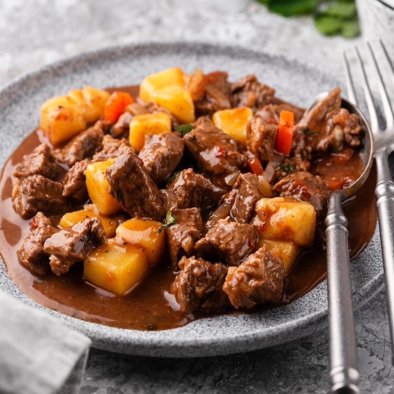 Hearty beef stew with potatoes and red bell peppers in a rich brown sauce on a gray plate.