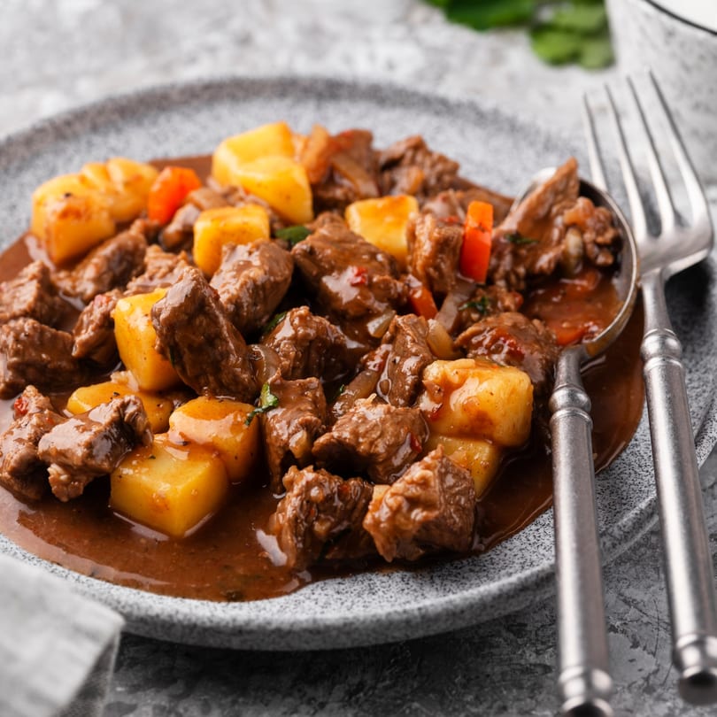 Hearty beef stew with potatoes and red peppers in a rich brown sauce, served on a gray plate.